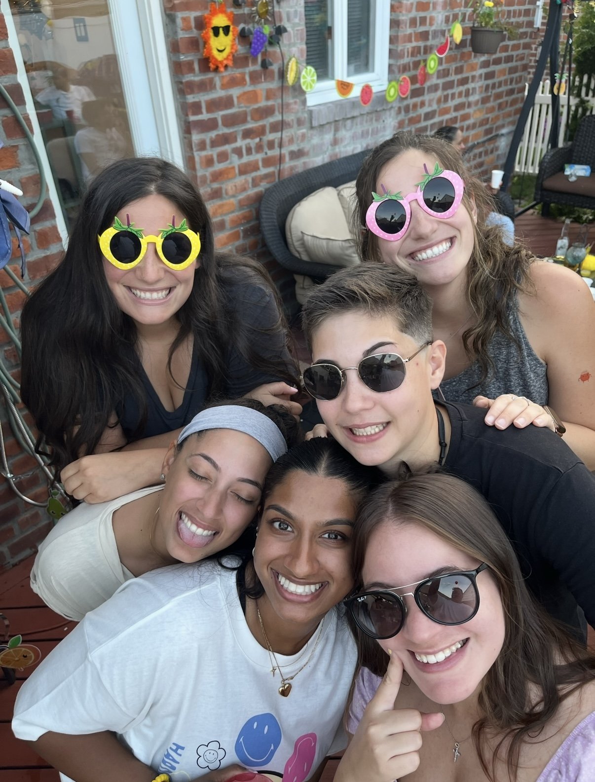 Six friends leaning in for a photo on a back deck wearing novelty fruit sunglasses